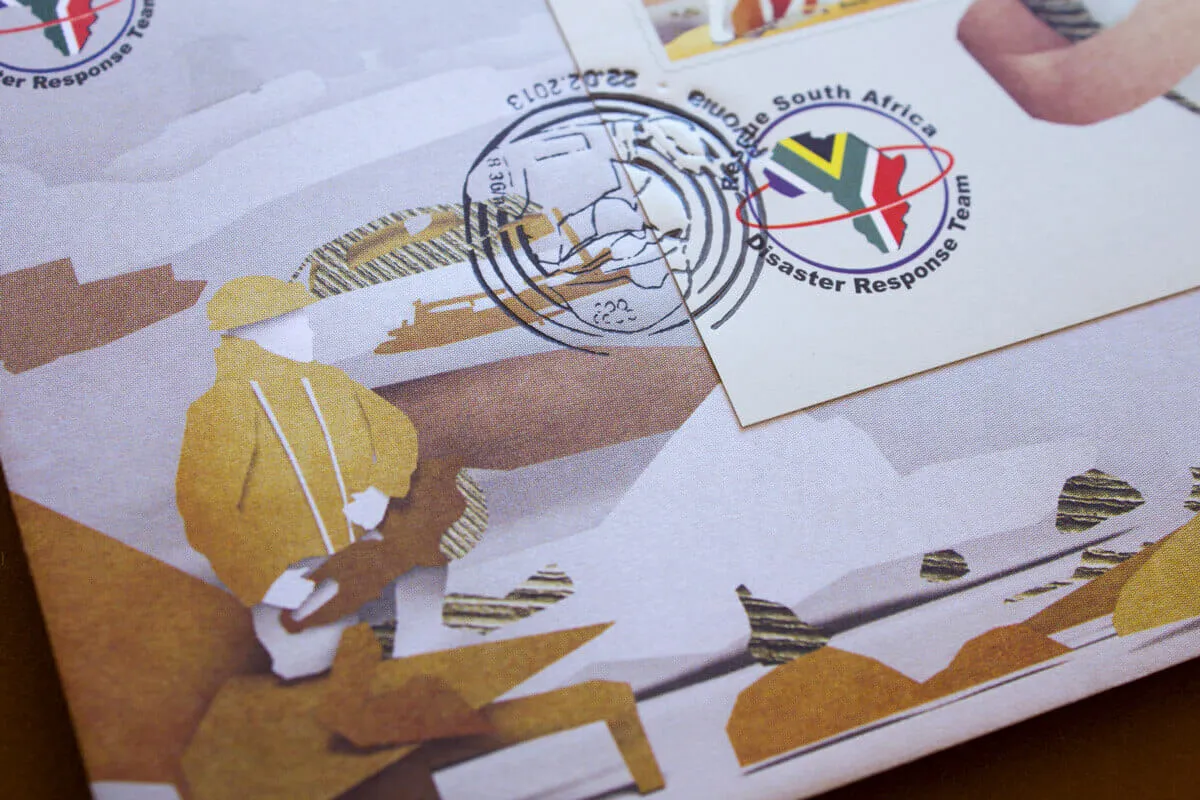 Wide detail of the envelope artwork. A kneeling figure in an ochre paper jacket works among broken slabs and corrugated debris, beside the Rescue South Africa emblem and a second cancellation.
